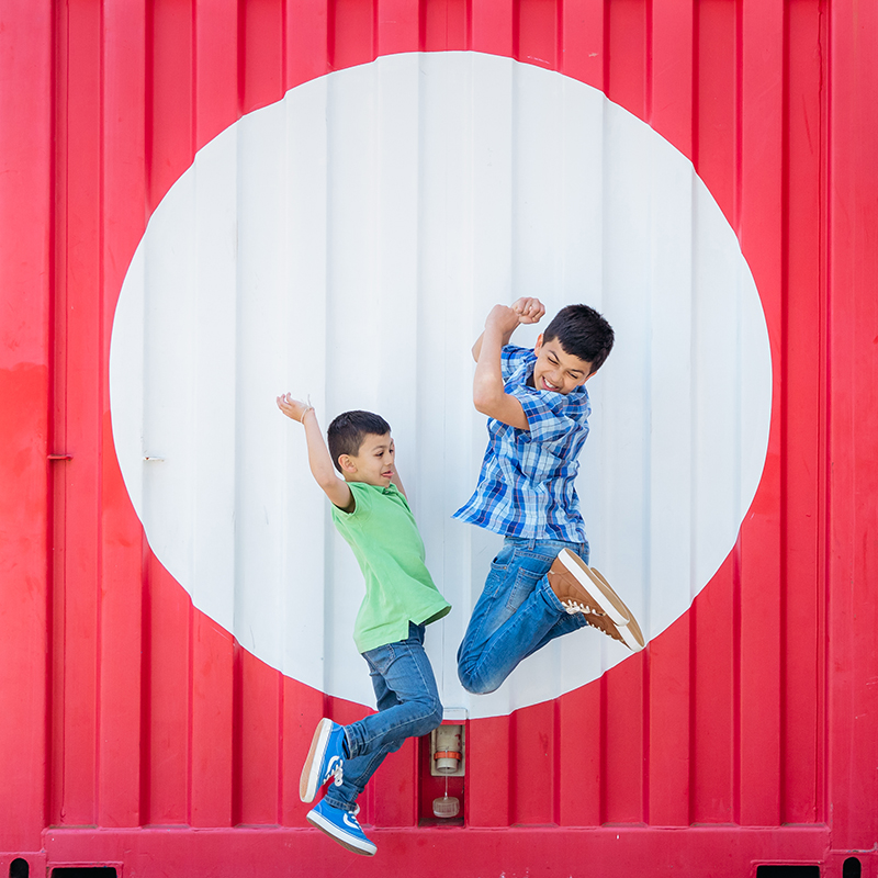 This is a fun, lively, vibrant and contemporary portrait of two boys. They are jumping up in the air in front of a red shipping container with a large red spot on it. The boys are jumping with their arms and legs up in the air with big smiles on their faces.
