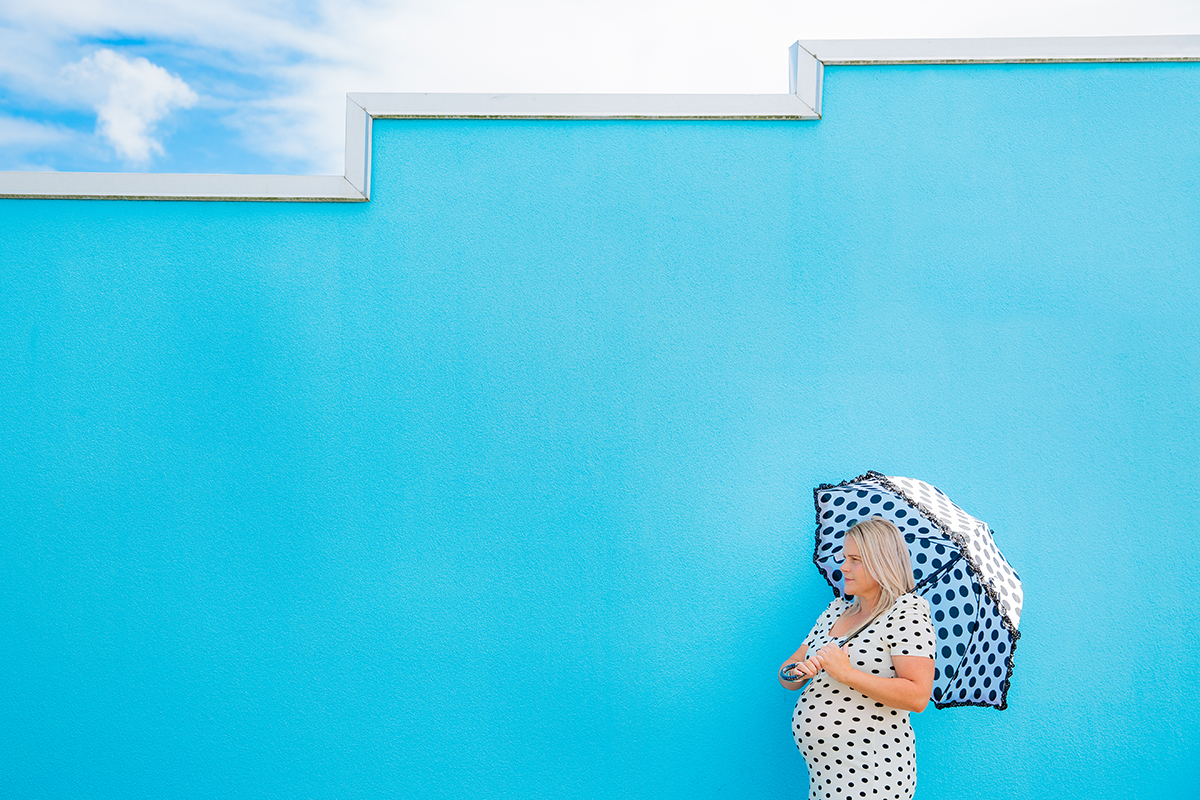 This is a photo taken during a maternity photo shoot in Auckland of a pregnant woman wearing a white and black polka dot dress. She is holding a black and white polka dot umbrella and is standing in front of a bright blue wall. Photo taken by Mala Photography, and Auckland based maternity photographer.