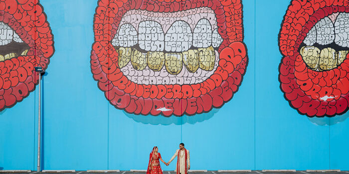 This is a photo from an Indian wedding in Christchurch, New Zealand. The photo is of a couple standing side by side holding hands in traditional Indian wedding outfits in front of a street art mural. The mural is of a bright red pair of lips on a bright blue background. Photo taken by Mala Photography, Auckland.
