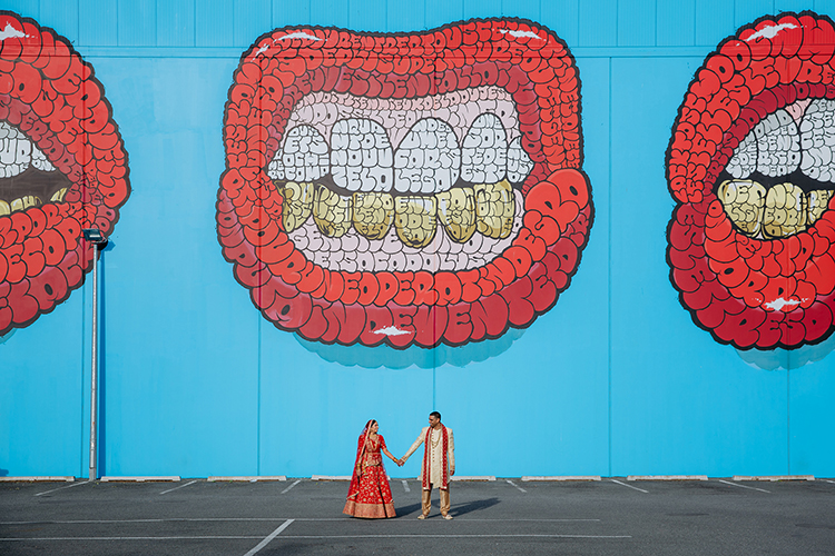 This is a photo from an Indian wedding in Christchurch, New Zealand. The photo is of a couple standing side by side holding hands in traditional Indian wedding outfits in front of a street art mural. The mural is of a bright red pair of lips on a bright blue background. Photo taken by Mala Photography, Auckland.