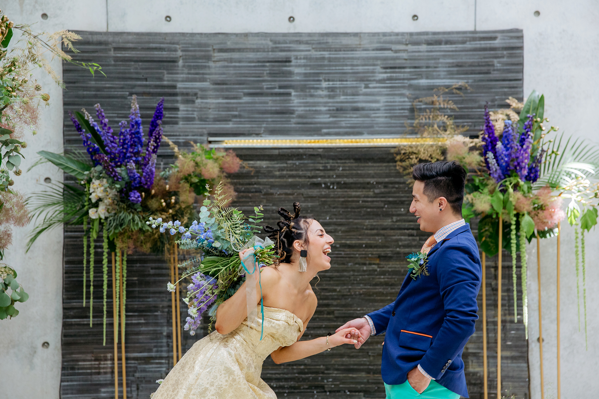 This is a photo of a Bride an Groom during their wedding ceremony at Cocoon House. They are looking at each other and laughing. The bride is dressed in a gold dress and she is holding a bouquet of flowers. The Groom is wearing a bright blue blazer, green pants and his holding her hand. They are laughing and joyful.
