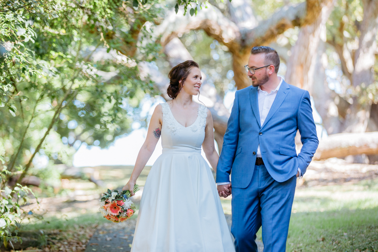 Mala Photography, a wedding photographer based in Auckland took this photo of a couple on their wedding day. They are walking on a path in a park, looking at each other, smiling and are hand in hand.
