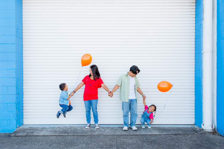 This is a family photo taken during a family portrait session by Mala Photography, and Auckland based photographer. This photo is of a family of four ie Mum, Dad and their son and daughter. They are playing with each other and having fun in front of a white roller door with bright blue brick work around it. The children are holding bright orange balloons and are jumping in the air.