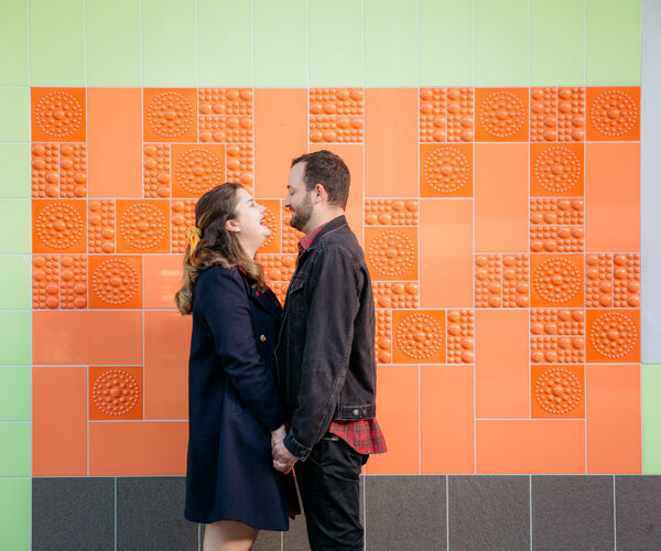 A photo from an inner city Auckland engagement shoot of a couple walking along holding hands and looking at each other. They are walking on a footpath with an orange, green and white tiled wall in the background. This photo was taken by Mala Photography. Mala is an Auckland based engagement and wedding photographer with a contemporary and creative style.