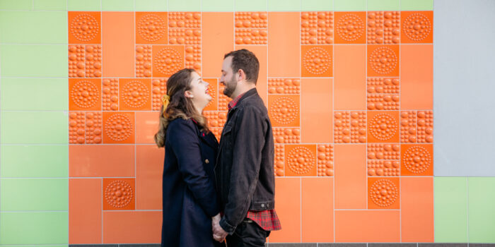 A photo from an inner city Auckland engagement shoot of a couple walking along holding hands and looking at each other. They are walking on a footpath with an orange, green and white tiled wall in the background. This photo was taken by Mala Photography. Mala is an Auckland based engagement and wedding photographer with a contemporary and creative style.