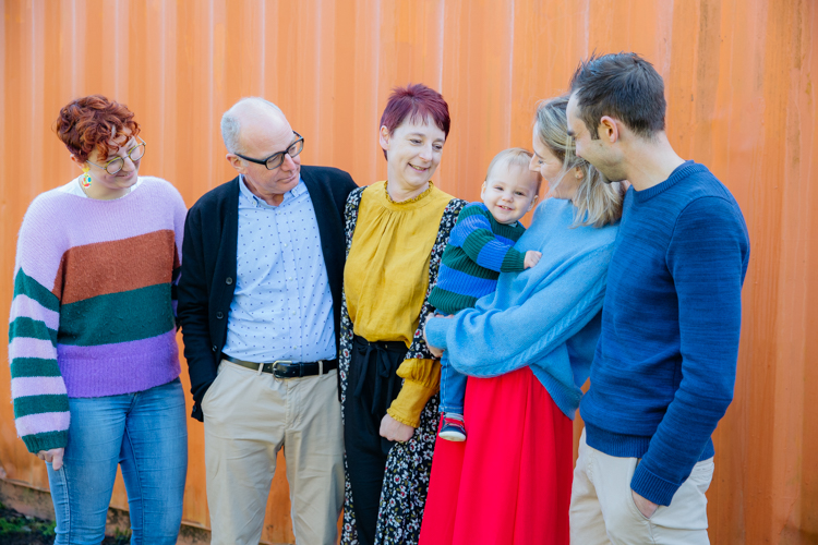 This is a family photo of six people - grandparents, parents an auntie and a one year old boy. They are standing in front of a bright orange shipping container smiling and laughing. The Mum is holding the little boy. He is looking at the camera with a very big smile on his face. This photo was taken by Auckland family photographer Mala Photography.