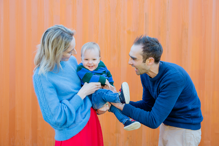 This is a photo of a family of three - Mum, Dad and their one year old son. The parents are holding the little boy and tickling him. He is looking at the camera and laughing. This photo was taken by Auckland based family portrait photographer Mala Photography.