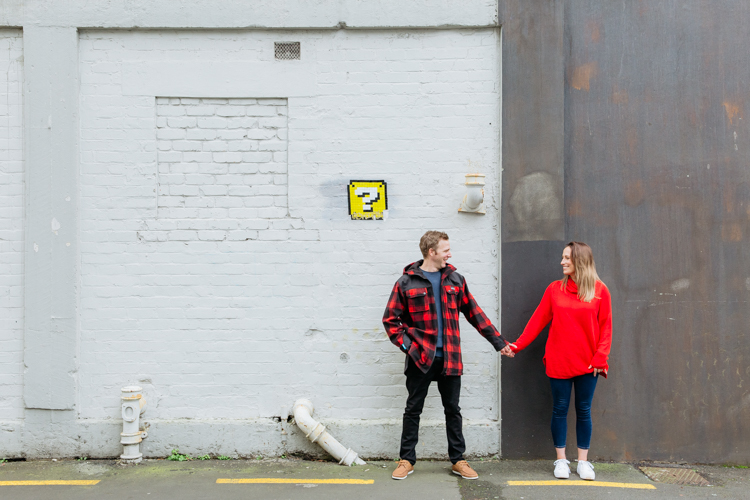 This is a photo of a newly engaged couple taken during their engagement shoot in Auckland. They are standing in front of a white brick wall in an alley way holding hands, looking at each other and smiling. They are dressed in bright red. This photo was taken by Mala Photography, an Auckland based wedding, engagement, portrait and event photographer.