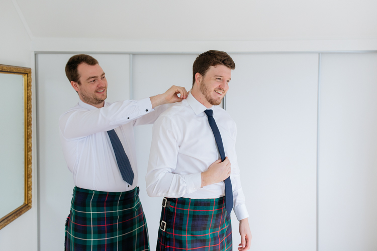 A groom wearing a kilt, shirt and tie getting ready on his wedding day. His groomsmen is helping him get ready and is adjusting his tie. Photo taken by a wedding photographer in Auckland, Mala Photography.